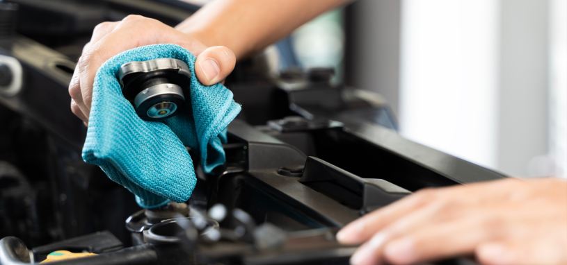 A closeup of a hand holding a radiator cap over a radiator. The hand is holding the cap with a small blue towel, just in case there’s any heat. However, the radiator is cool.