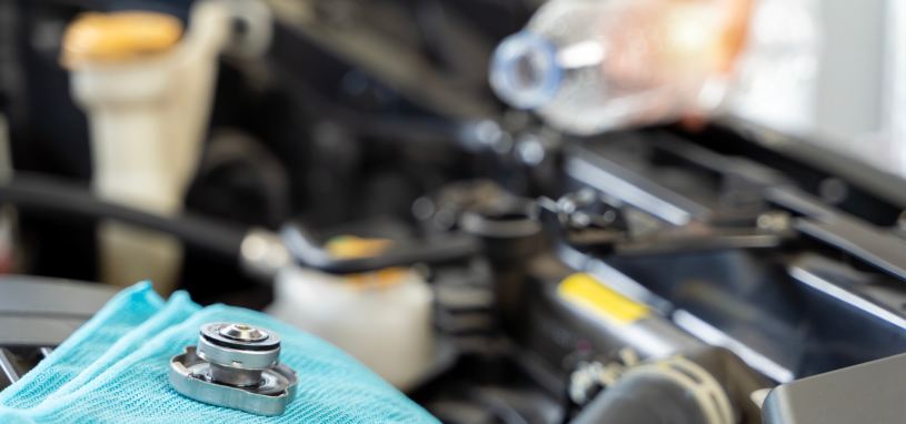 A car owner has safely removed the cool radiator cap with a towel and left it to the side. They are now refilling their radiator with distilled water from a bottle as a temporary solution till they can reach a radiator mechanic.
