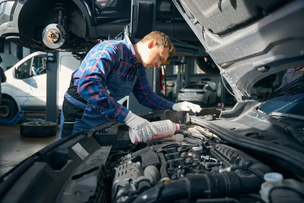 A mechanic wearing a blue plaid shirt, overalls, and gloves pours coolant into a car engine while working in a professional auto repair shop.