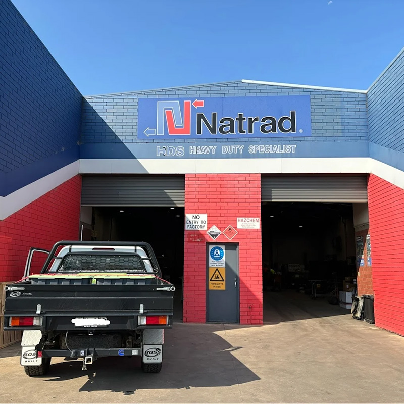 A blue and red Natrad auto repair workshop with a black pickup truck parked in front of two large open garage bay doors.