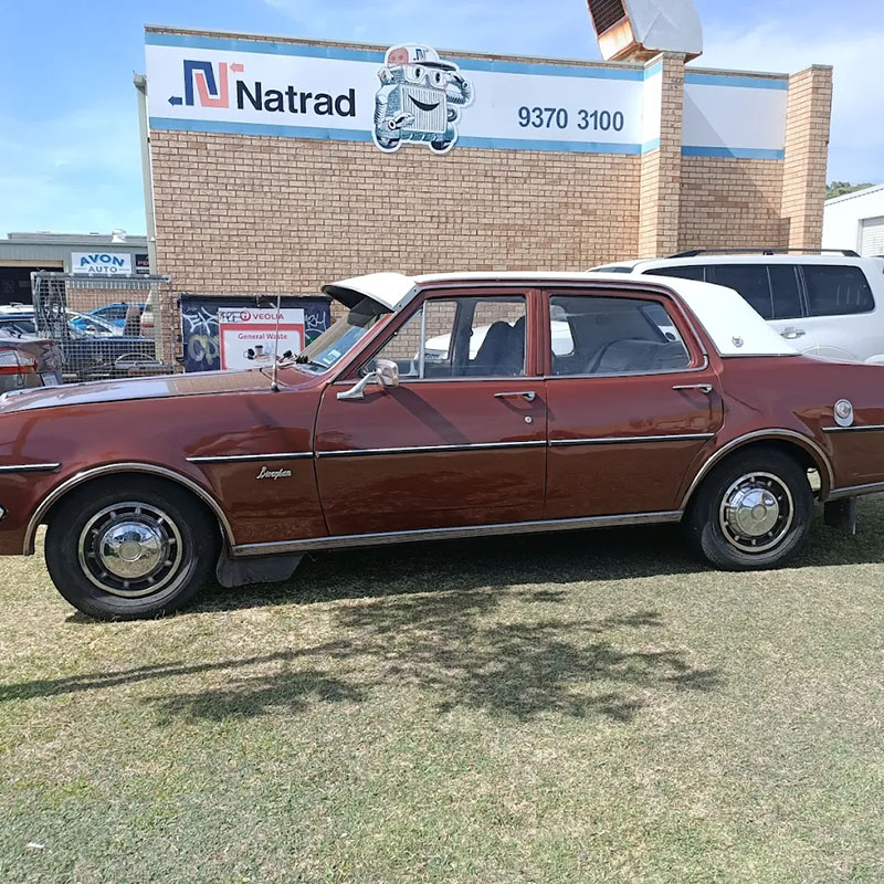 A classic brown sedan with a white roof parked on the grass in front of a Natrad Radiators & Auto Air service centre.