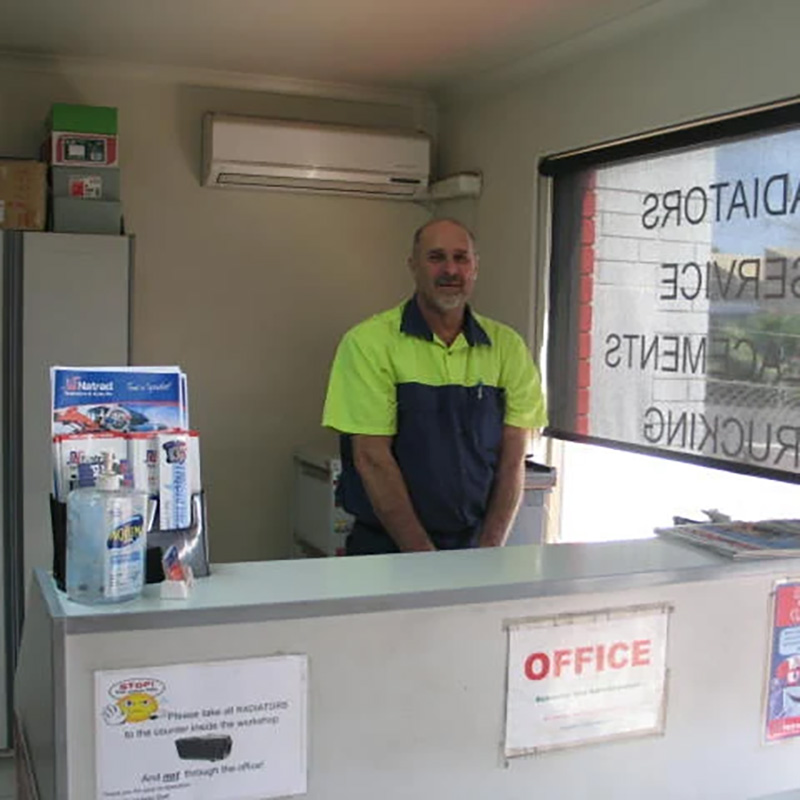 A mechanic in a high-visibility shirt stands behind a service counter in a bright office at an auto radiator repair shop.