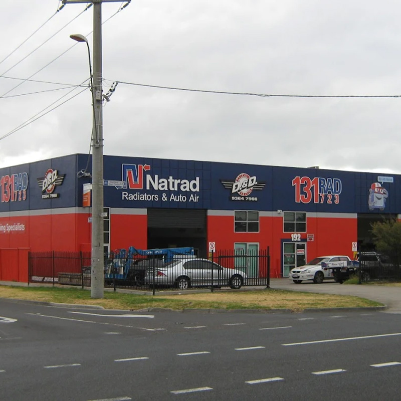 Large red and blue Natrad Radiators & Auto Air workshop on a street corner with parked cars and a cloudy sky above.