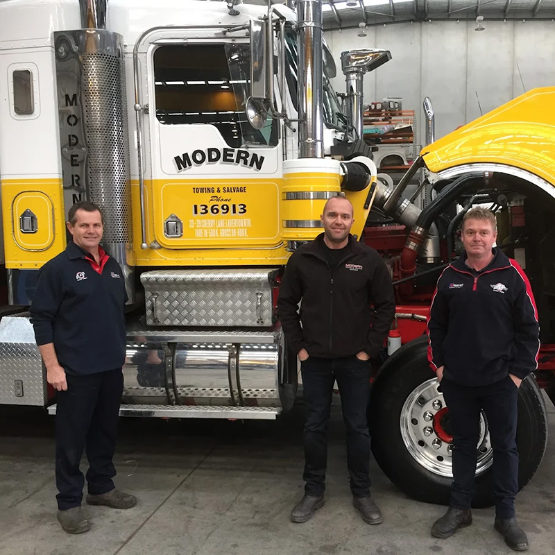 Three mechanics stand in front of a large white and yellow semi-truck with its hood open in a repair workshop.