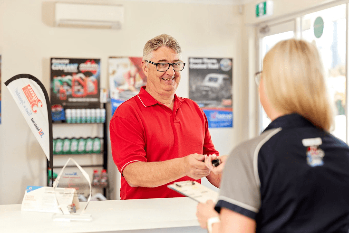 A smiling customer in a red shirt hands car keys to a Natrad staff member at a service counter in a bright office.