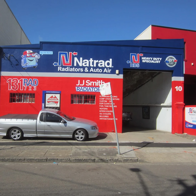 A blue and red Natrad workshop with JJ Smith signage, a silver ute parked in front, and a large open garage bay door.