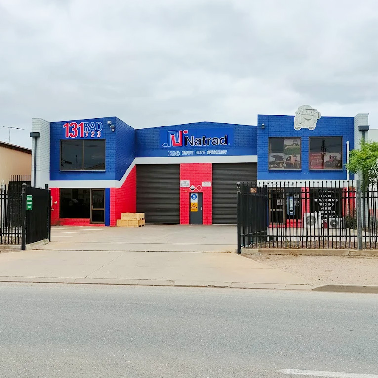 A blue and red Natrad auto repair building with closed garage bays, a fenced yard, and signage under a cloudy sky.
