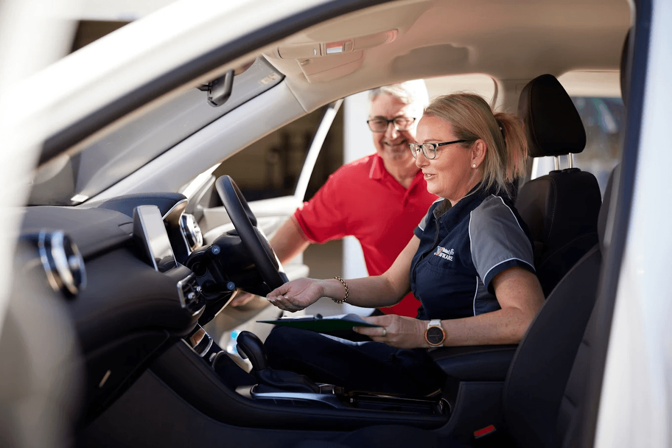 A female technician in a Natrad uniform sits in a car, discussing a clipboard with a smiling customer in a red shirt.
